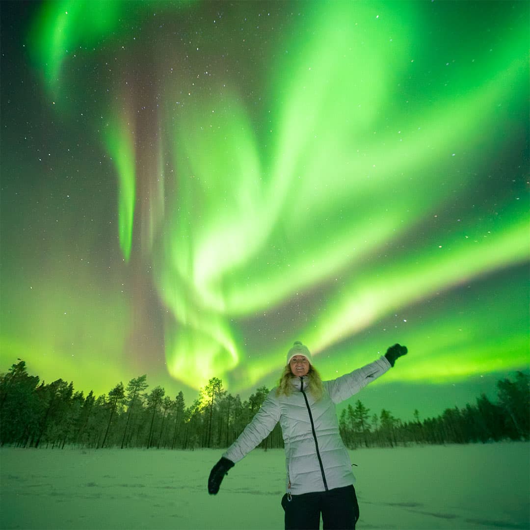 A guest with arms raised under swirling northern lights on a snowy open field