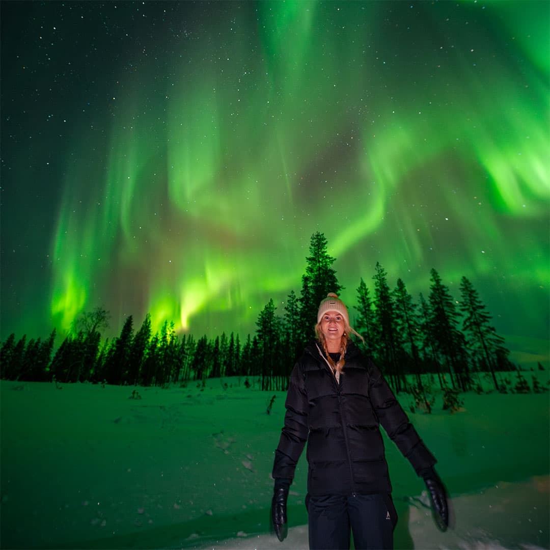 A guest in a winter jacket under bright green northern lights in a snowy forest clearing