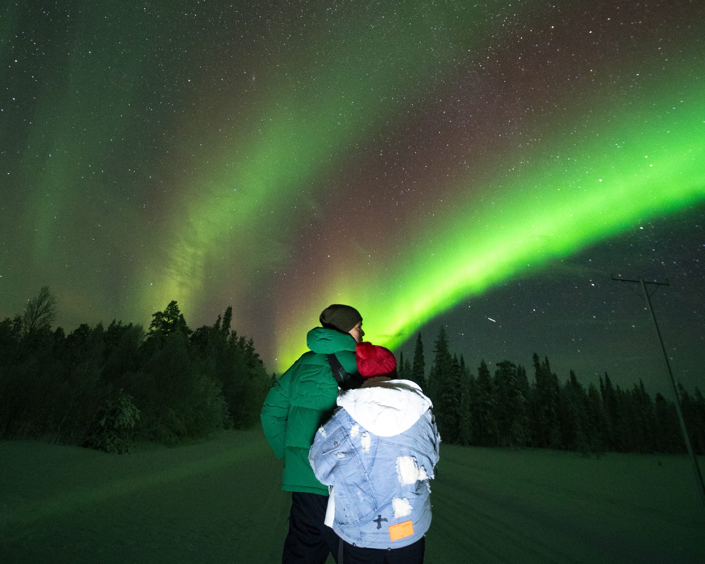 Two guests side by side watching a band of northern lights over a snowy forest